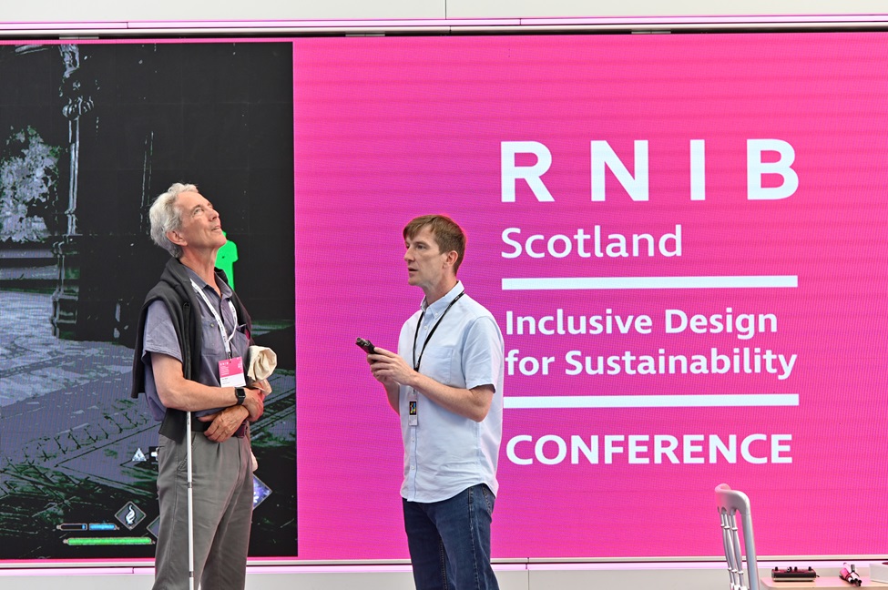 Barry Snell and Ken Reid from RNIB Scotland chat next to a large logo of the Conference